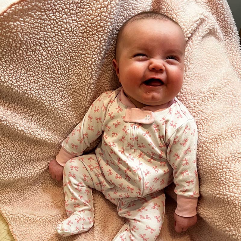 Smiling baby wearing pink bow-patterned pajamas, lying on a soft pink blanket.