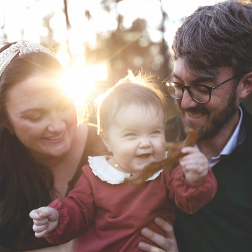 Family-Building-Grants_HeaderImage Smiling mother and father holding their laughing baby outdoors, with warm sunlight shining behind them.