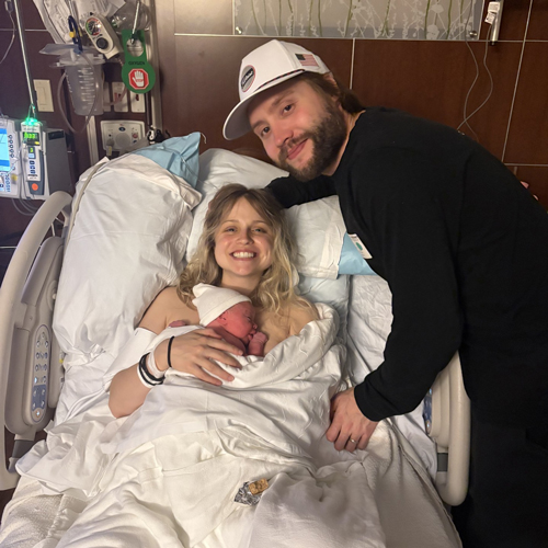 Smiling mother holding her newborn baby in a hospital bed, with the father leaning beside them.