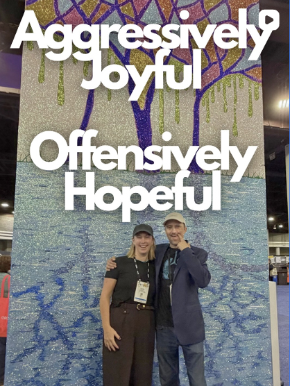 Two people smiling together in front of a colorful display featuring the words “Aggressively Joyful” and “Offensively Hopeful.”