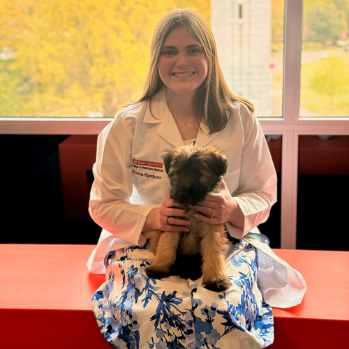 Impact_Anna-Spencer-WCC-with-Sulley Smiling woman in a white medical coat holding a small brown and black puppy, with colorful autumn foliage visible through the window behind her.