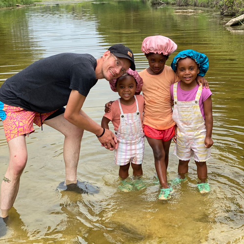 Impact_Casey-McPhail-with-daughters Smiling man and three young girls standing together in shallow river water, wearing colorful swimwear and bonnets.
