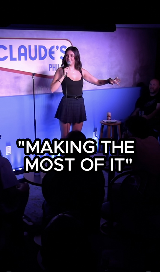 Kristen Cills performing stand-up comedy onstage at a comedy club, holding a microphone and gesturing toward the audience.