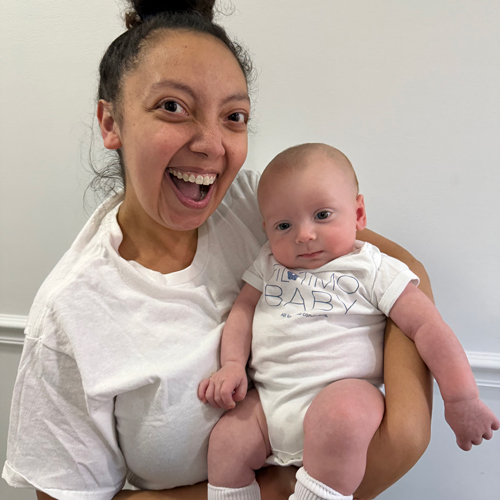 Smiling woman holding a baby wearing a white Filotimo Baby onesie.