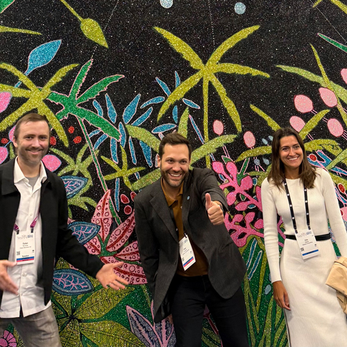 Dylan, Pete, and Ella posing in front of a colorful tropical mural, with Pete giving a thumbs-up.