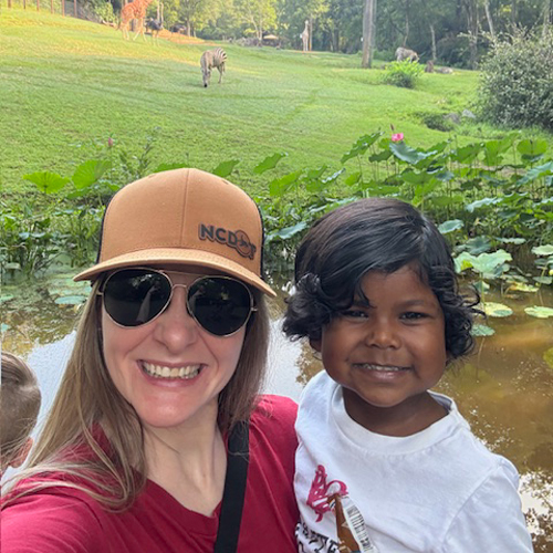 WhatWeCover_Anna-and-Divya-Ward_Adoption Smiling woman wearing sunglasses and a baseball cap posing with a young child beside a pond, with zebras grazing in the background.