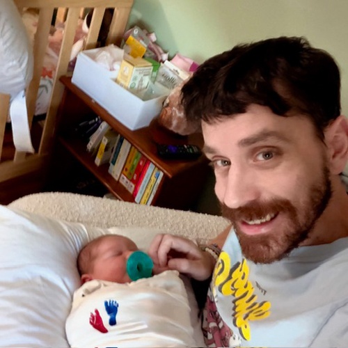 Who-Can-Apply_Lilian_Brian_Donahoe Smiling father sitting beside his newborn baby, who is wrapped in a blanket with a pacifier.